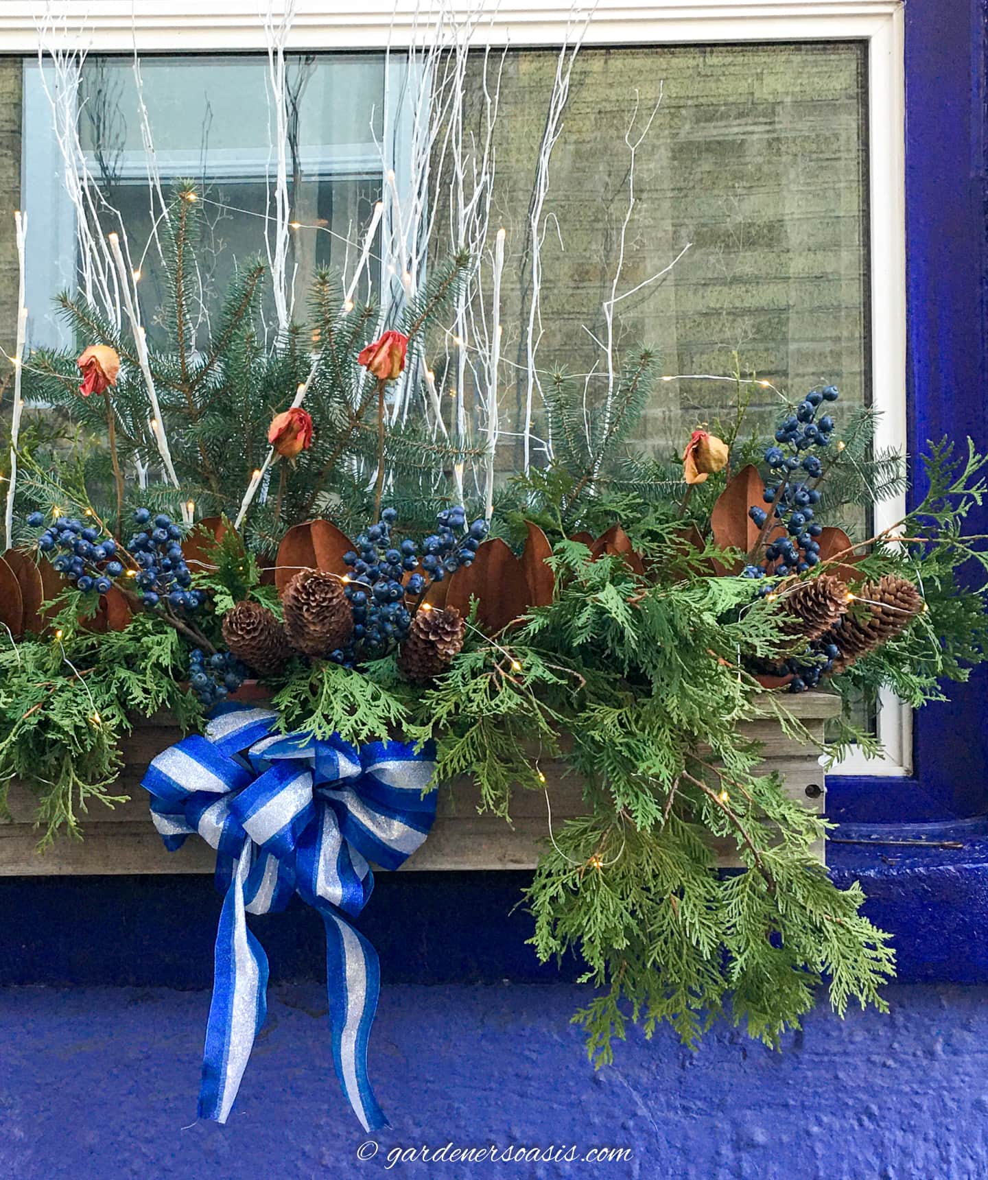 An evergreen window box with pine cones, white twigs and a blue and white bow
