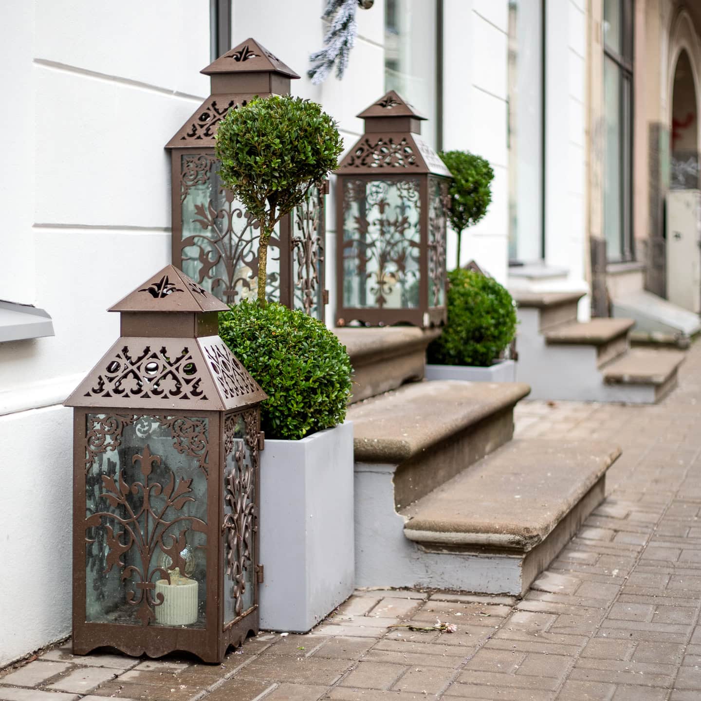Large ornate metal lanterns and round topiary plants in square pots are arranged on and beside outdoor stone steps leading to a building entrance.