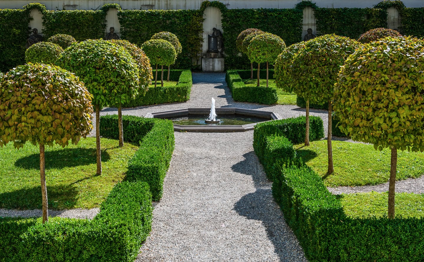 Formal garden with neatly trimmed hedges, round topiary trees, gravel pathways, and a central fountain surrounded by lush greenery.