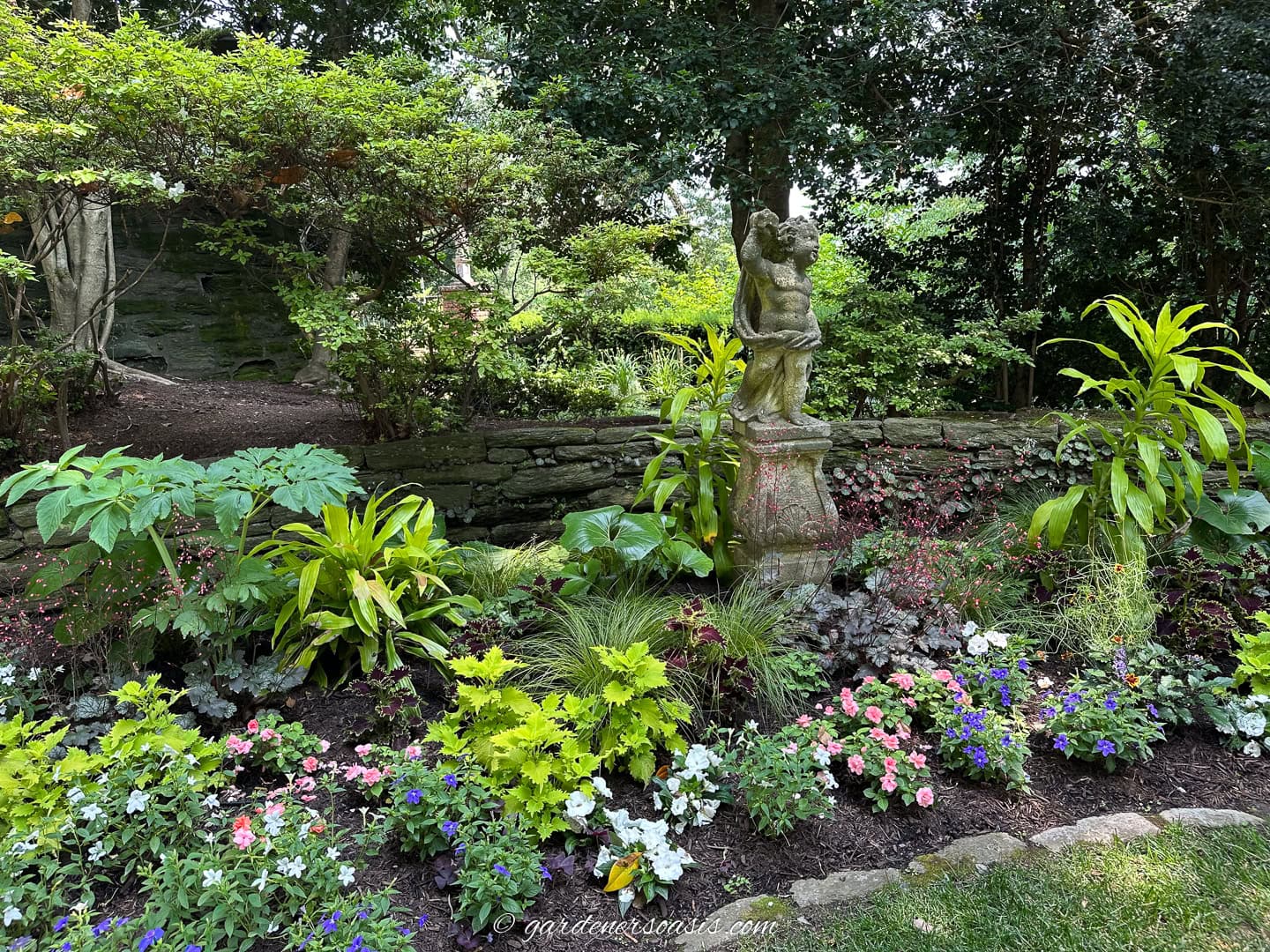 A stone statue stands in a lush garden bed, surrounded by flowering plants and greenery, with a stone wall and trees in the background.