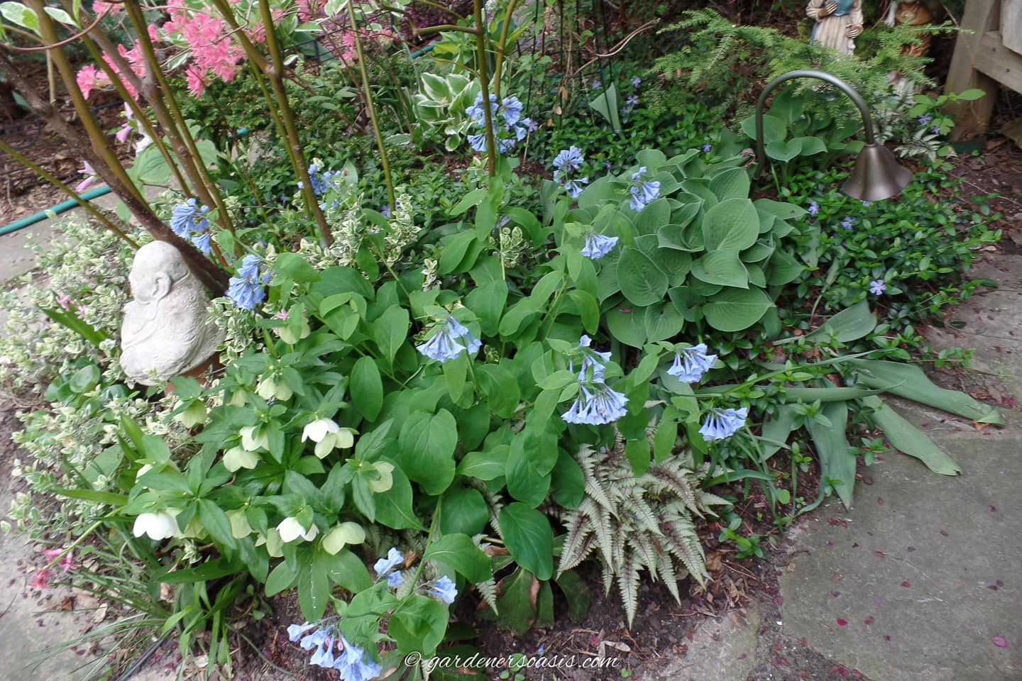 A garden bed with green foliage, blue and white flowers, a small garden statue, and a metal light fixture beside a stone pathway.