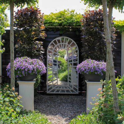 a large white garden mirror on a black fence with symmetrical plantings around it